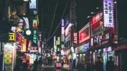 Sebuah jalan yang hidup di Seoul di malam hari dengan papan iklan neon dan orang-orang yang berjalan, menangkap esensi kehidupan perkotaan di Korea Selatan.