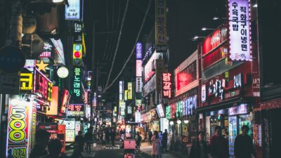 Sebuah jalan yang hidup di Seoul di malam hari dengan papan iklan neon dan orang-orang yang berjalan, menangkap esensi kehidupan perkotaan di Korea Selatan.