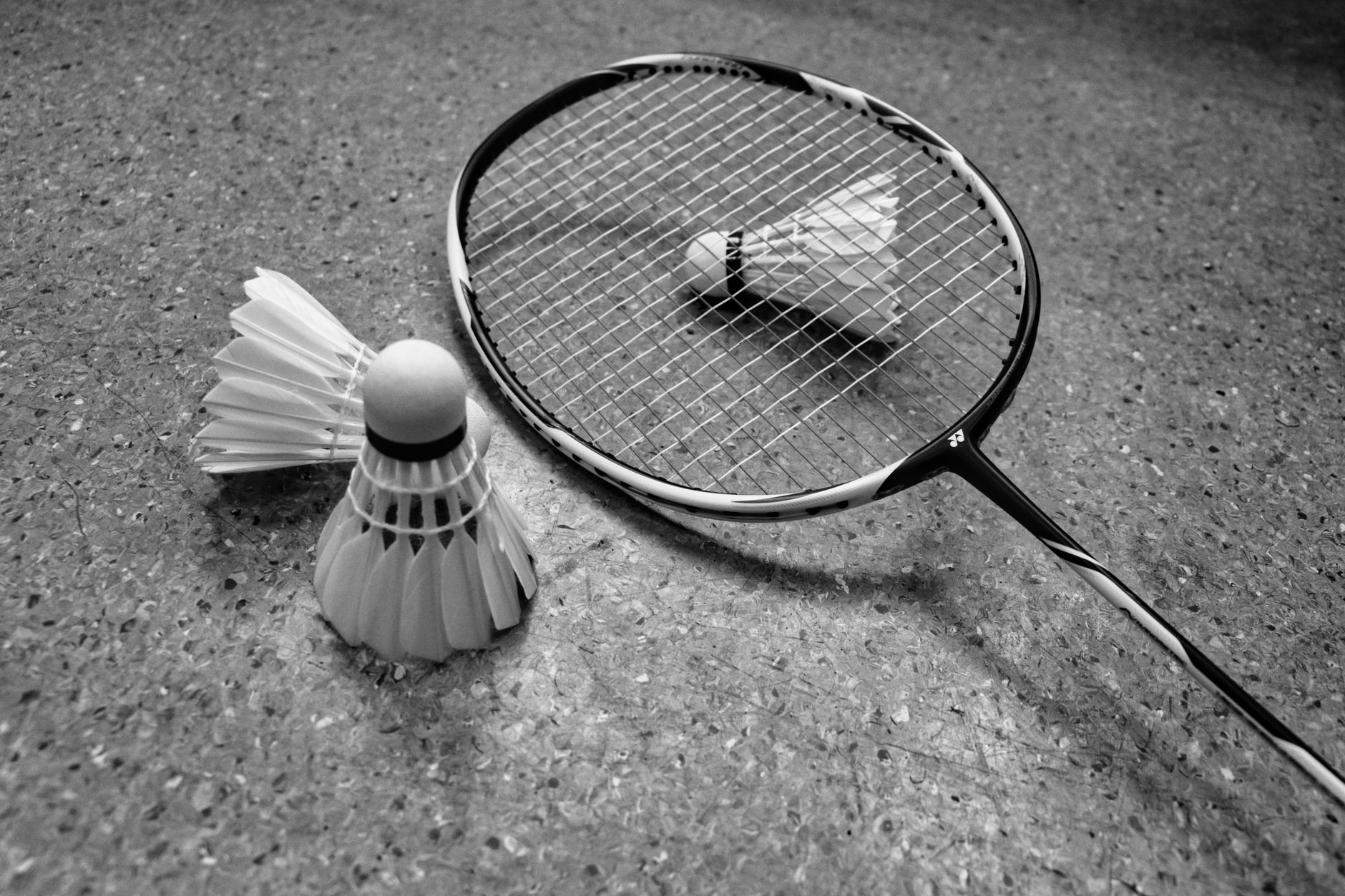 Gambar grayscale dari raket badminton dan kok di permukaan lapangan.