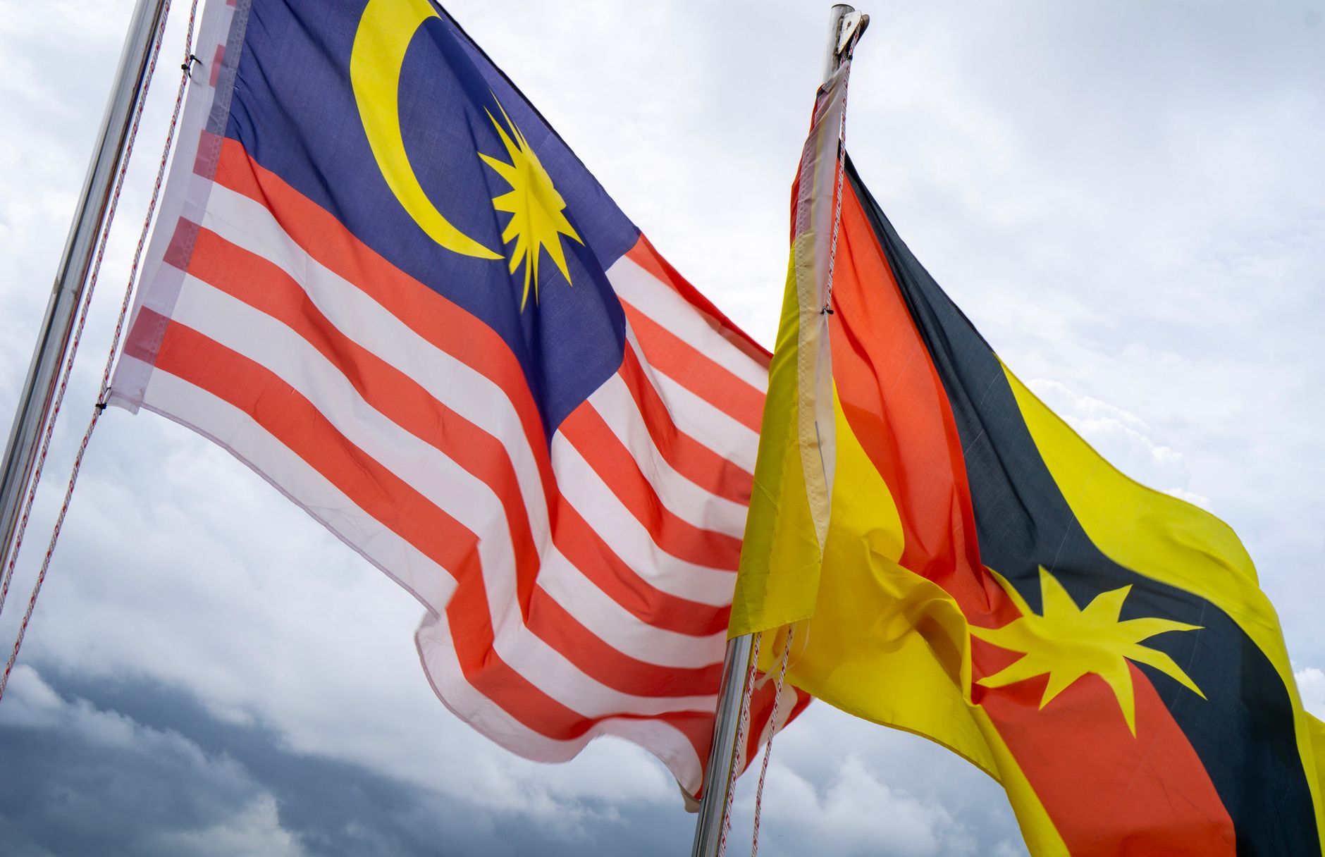 Bendera Malaysia dan Sarawak berkibar bersama di bawah langit yang mendung, melambangkan persatuan dan kebanggaan.