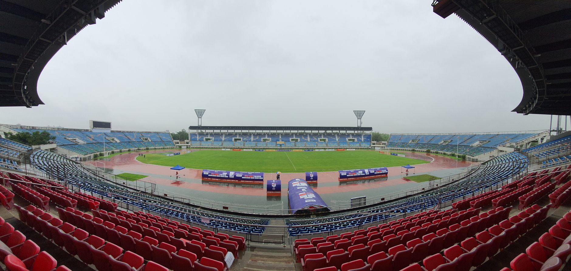 Pengambilan sudut lebar dari stadion olahraga kosong di bawah langit kelabu, menonjolkan area tempat duduk merah dan biru.