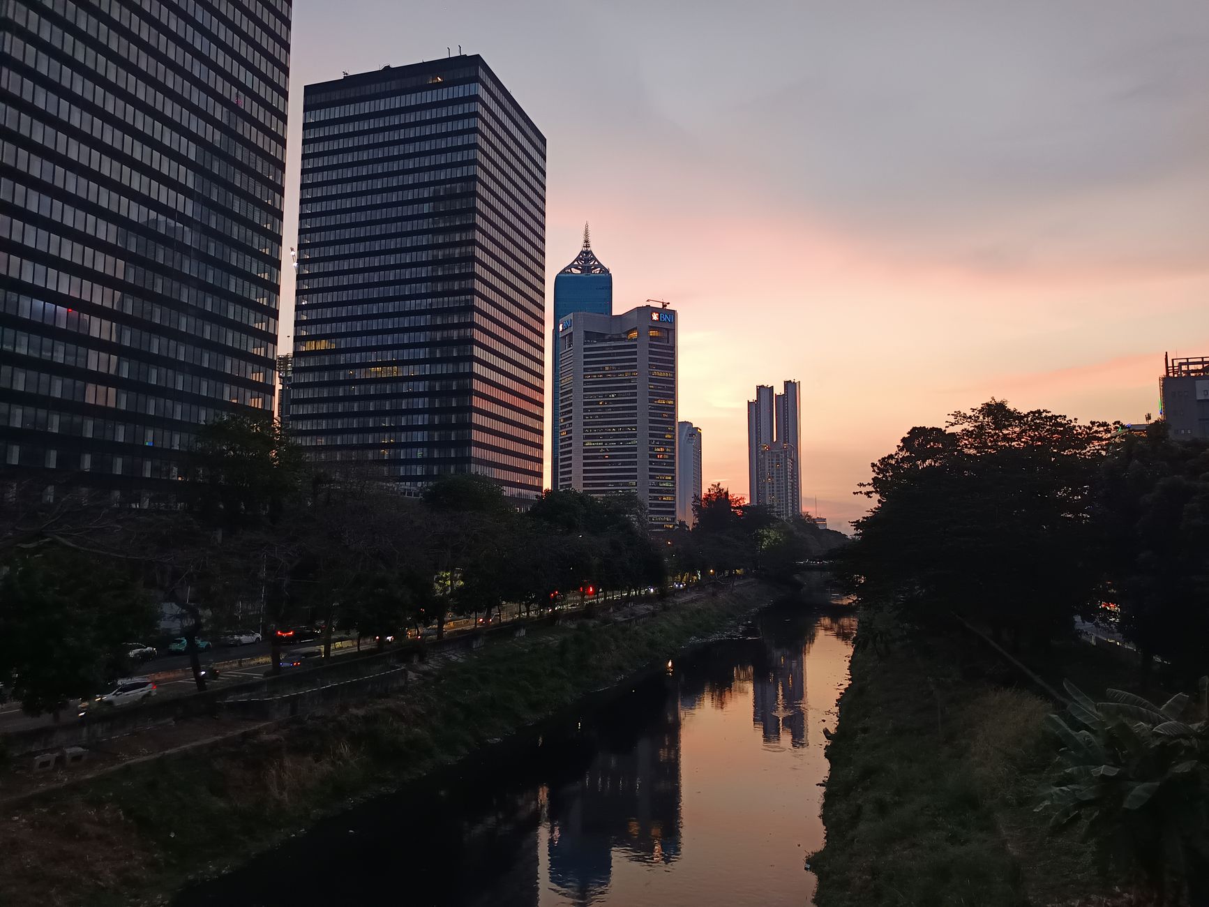 Pemandangan menakjubkan cakrawala Jakarta saat matahari terbenam, terpantul di kanal, menampilkan arsitektur modern.