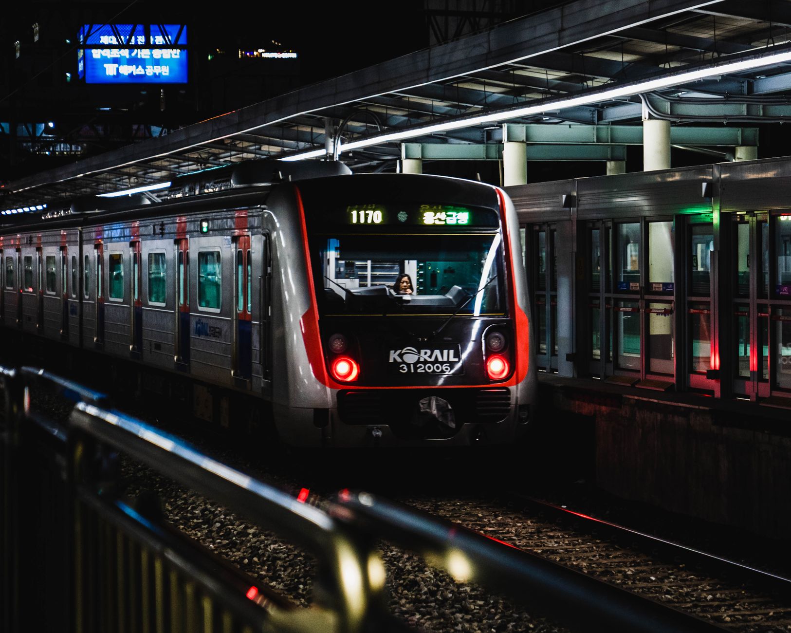 Sebuah kereta Korail di malam hari di stasiun Seoul yang ramai, disinari lampu-lampu yang cerah.