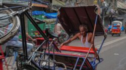 Seorang pria tua mengangkat rickshaw di jalan yang sibuk di Delhi, India, menunjukkan kehidupan jalanan yang cerah.