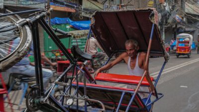 Seorang pria tua mengangkat rickshaw di jalan yang sibuk di Delhi, India, menunjukkan kehidupan jalanan yang cerah.