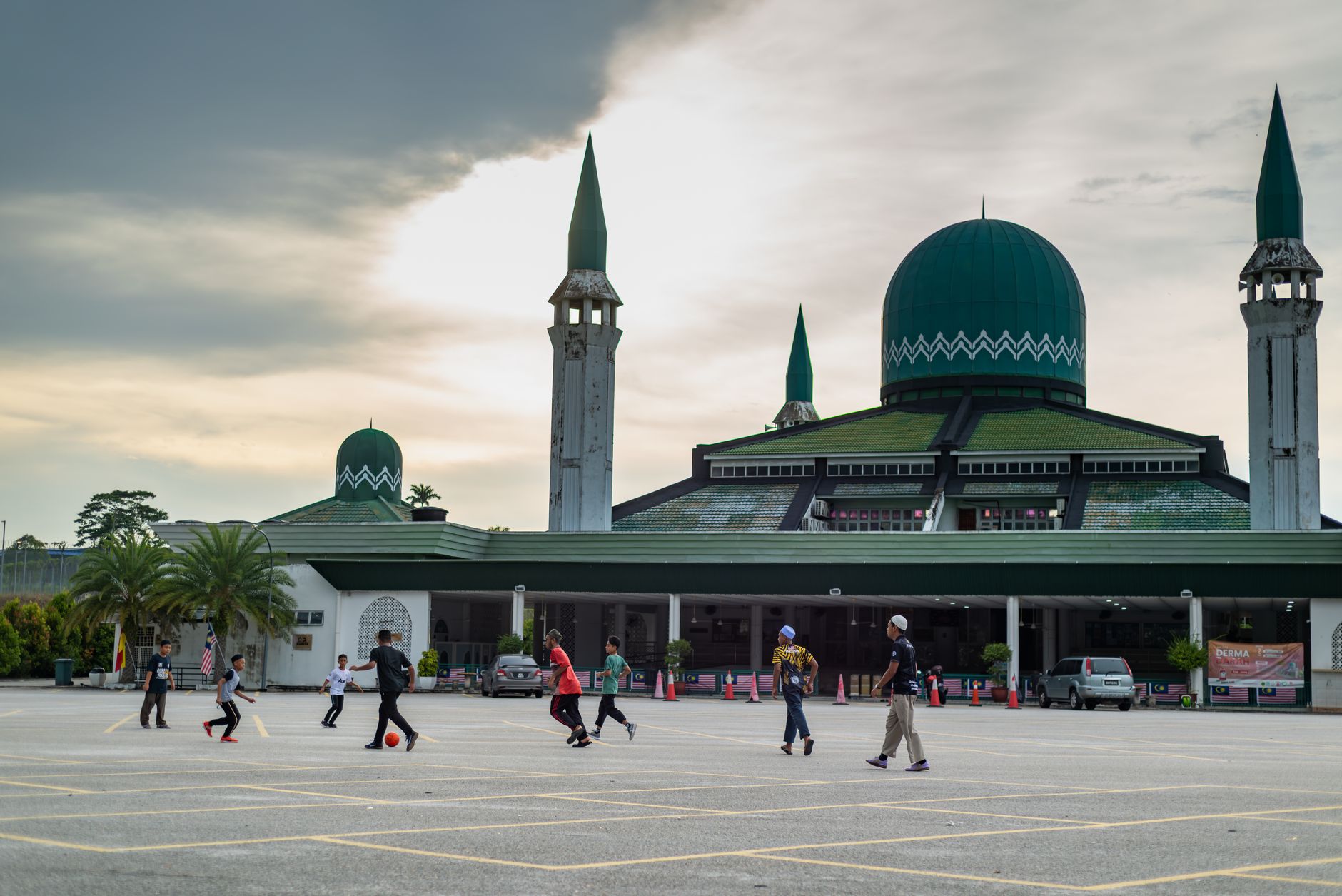 Anak-anak bermain sepak bola di depan masjid dengan kubah hijau dan menara, di bawah langit yang mendung.