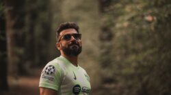 Seorang pria berjanggut dalam jersey sepak bola berdiri dengan percaya diri di lingkungan hutan yang tenang.