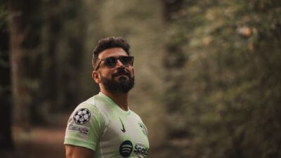 Seorang pria berjanggut dalam jersey sepak bola berdiri dengan percaya diri di lingkungan hutan yang tenang.