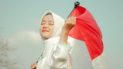 Wanita tersenyum mengenakan hijab, dengan bangga memegang bendera Indonesia di luar ruangan.