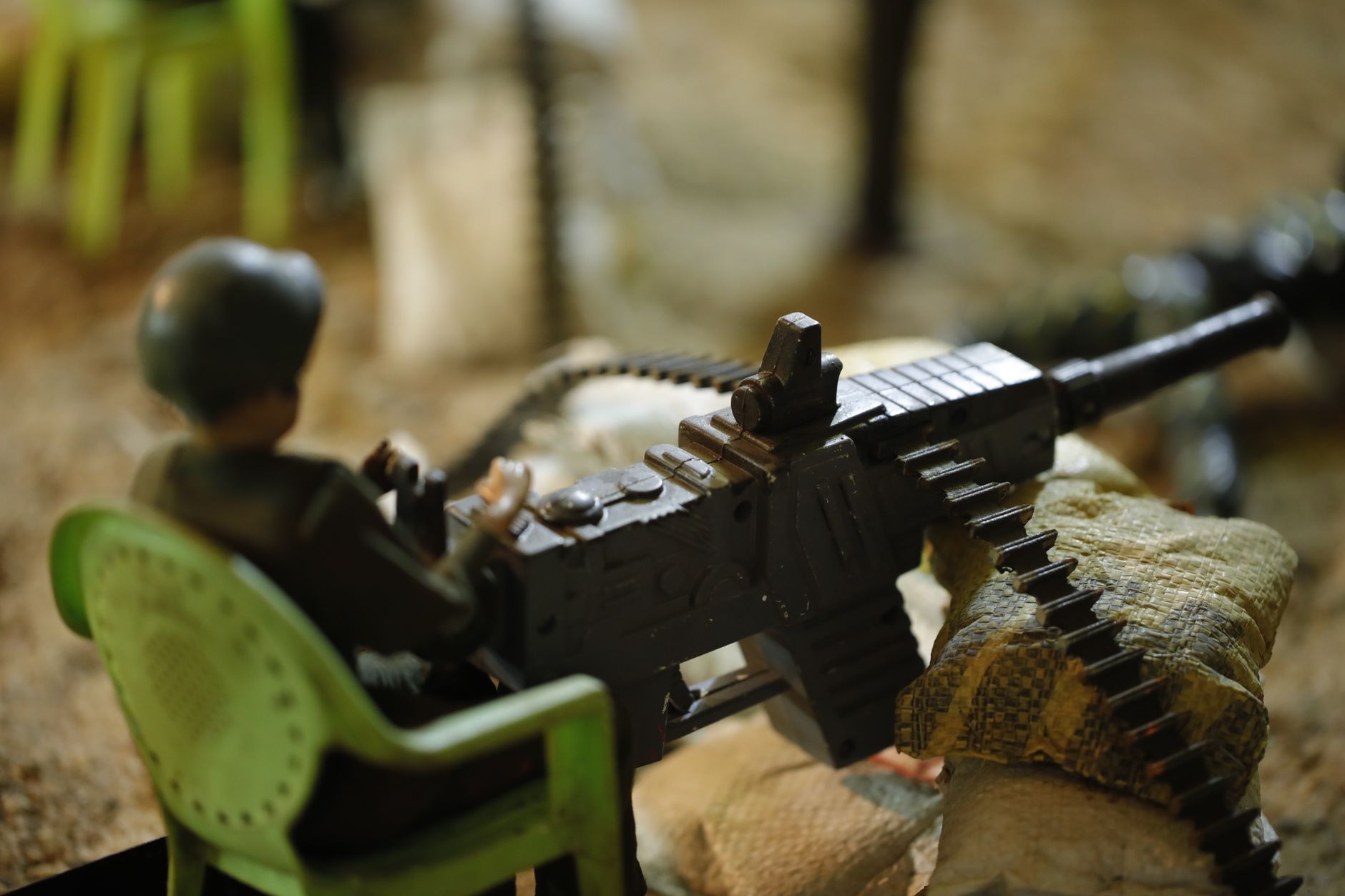 Close-up tentara mainan dalam pengaturan diorama, memegang senapan mesin plastik dan duduk di kursi hijau.