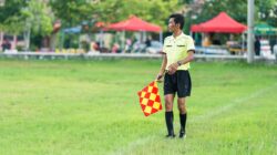 Update: Prosedur Pengamanan, Suporter Timnas Malaysia Keluar Paling Akhir dari GBK 2 Seorang hakim garis sepak bola berseragam berdiri di lapangan berumput dengan bendera selama pertandingan siang hari.