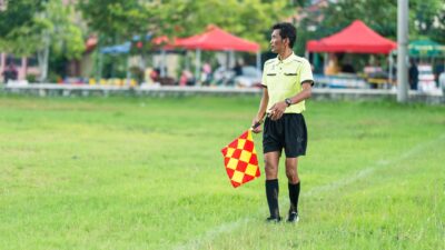 Update: Prosedur Pengamanan, Suporter Timnas Malaysia Keluar Paling Akhir dari GBK 8 Seorang hakim garis sepak bola berseragam berdiri di lapangan berumput dengan bendera selama pertandingan siang hari.