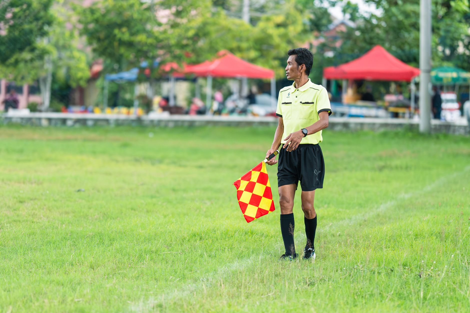 Seorang hakim garis sepak bola berseragam berdiri di lapangan berumput dengan bendera selama pertandingan siang hari.