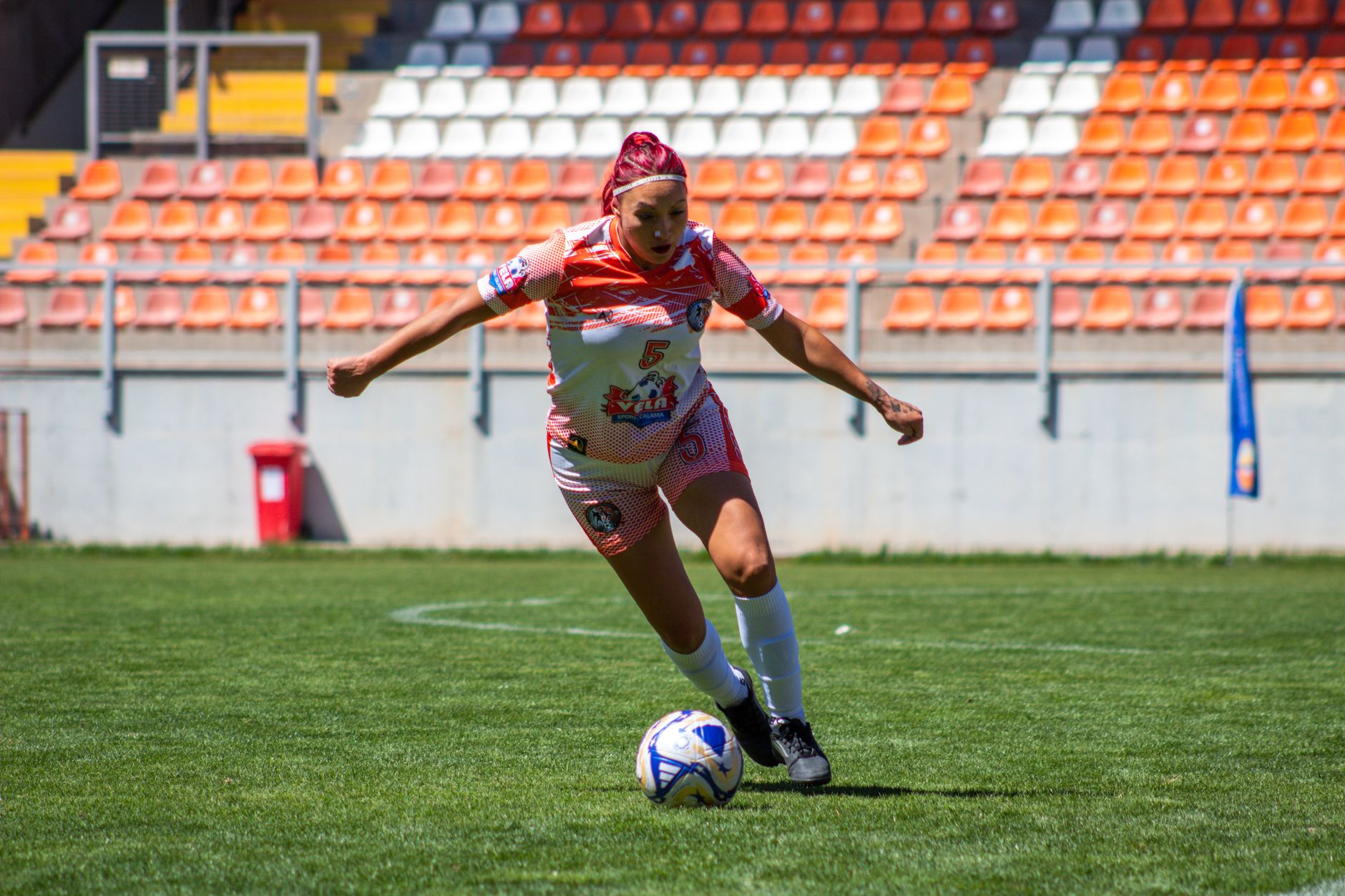 Seorang pemain sepak bola wanita dengan terampil menggiring bola di lapangan stadion luar ruangan yang cerah.