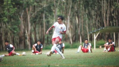 Pemain sepak bola yang dinamis dalam aksi selama sesi pelatihan luar ruangan dengan rekan tim di latar belakang.