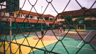 Orang-orang bermain di lapangan basket luar ruangan yang dikelilingi oleh pagar jaring.