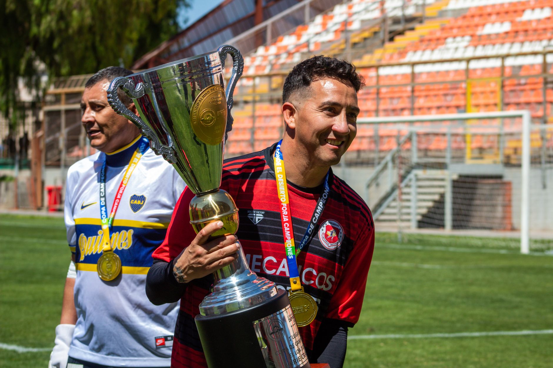 Dua pemain sepak bola merayakan dengan piala kejuaraan di lapangan.