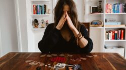 Seorang wanita yang sedang berpikir dalam-dalam duduk di meja dengan manik-manik dan sebuah foto, mengisyaratkan introspeksi.