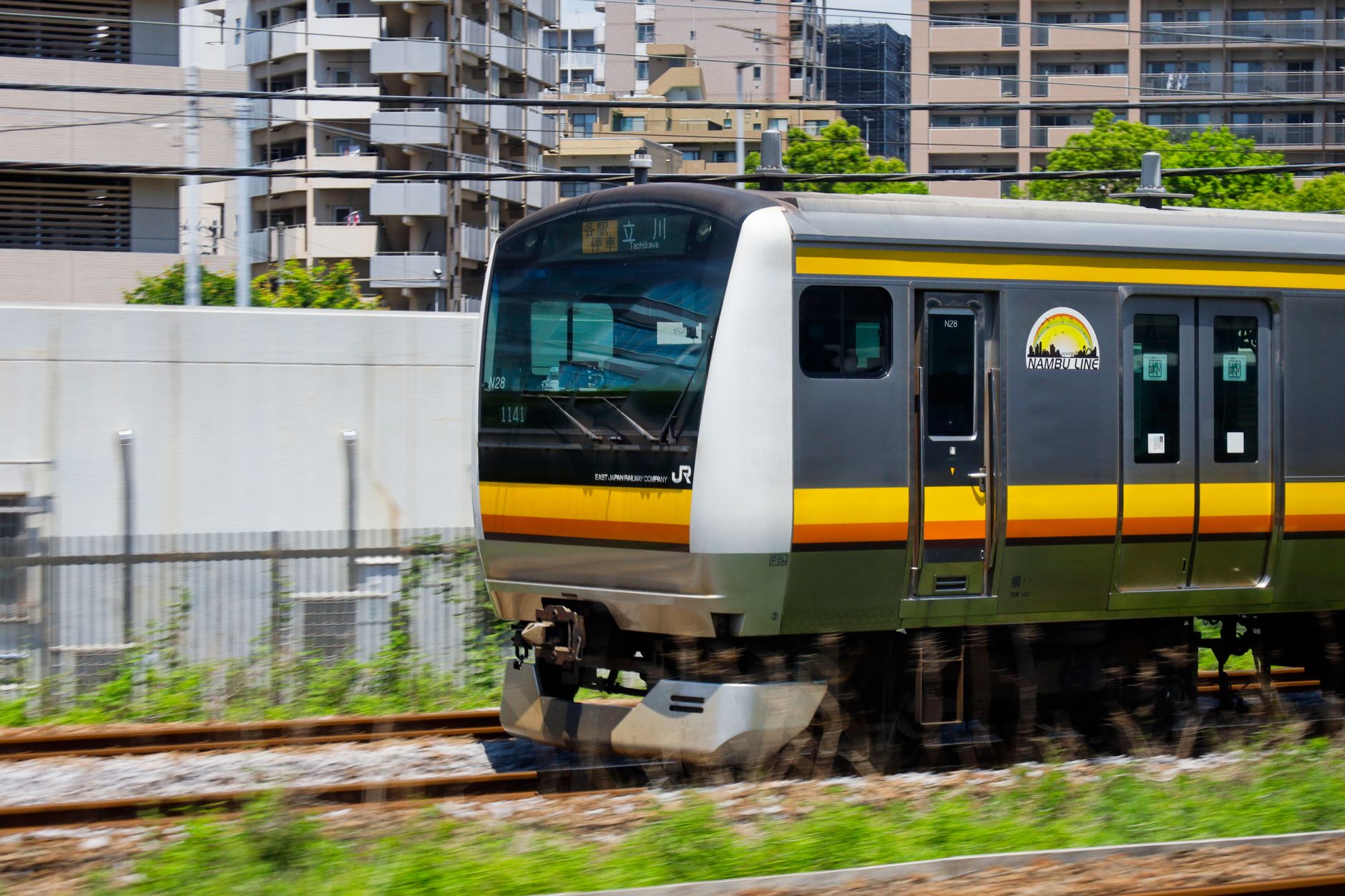 Kereta seri E233 di Jalur Nambu melintas di perkotaan Tokyo pada siang hari.