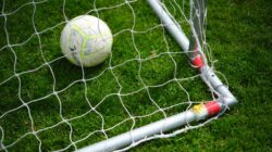 Gambar close-up sebuah bola sepak yang berada di dalam jaring gawang di lapangan rumput hijau yang cerah.