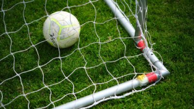 Gambar close-up sebuah bola sepak yang berada di dalam jaring gawang di lapangan rumput hijau yang cerah.