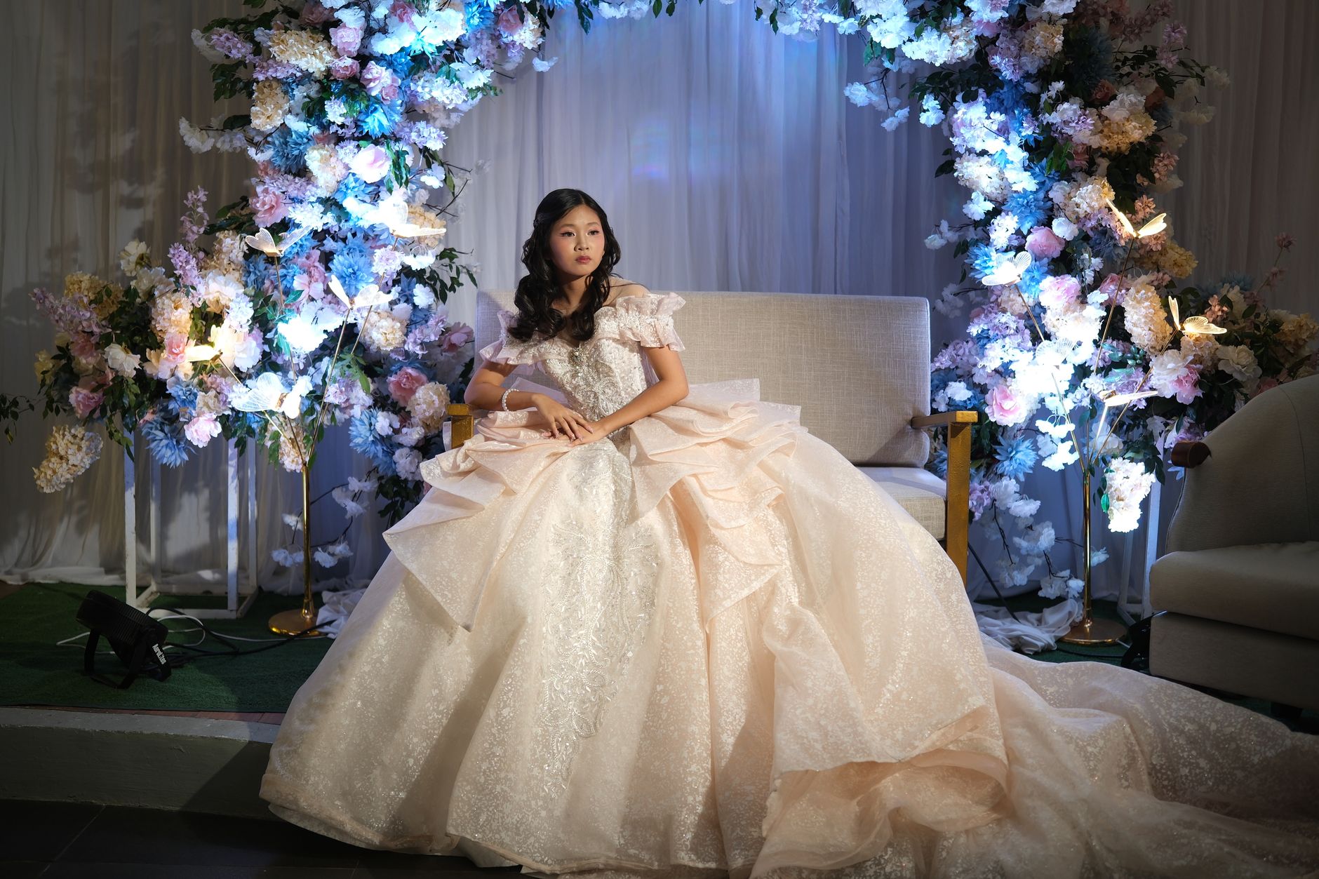 Debutante cantik dalam gaun yang dirancang dengan rumit, duduk di antara rangkaian bunga di acara formal.