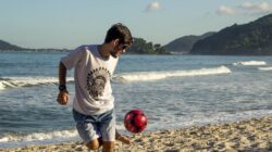 Seorang pria muda menikmati bermain sepak bola di pantai yang cerah dengan ombak dan pegunungan di latar belakang.