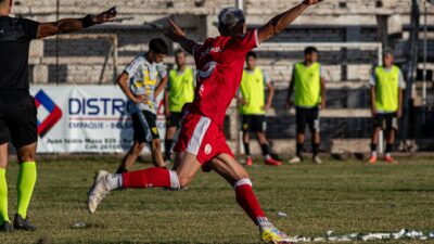 Seorang pemain sepak bola berdasi merah merayakan gol dengan antusias di lapangan luar ruangan.