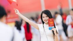 Sorotan: Sold Out! 67 Ribu Tiket Final FIFA ASEAN Cup 2026 Timnas Indonesia Vs Thailand Ludes Terju... 3 Seorang wanita tersenyum mengenakan gaun putih melambaikan bendera merah di sebuah acara luar ruangan yang meriah.