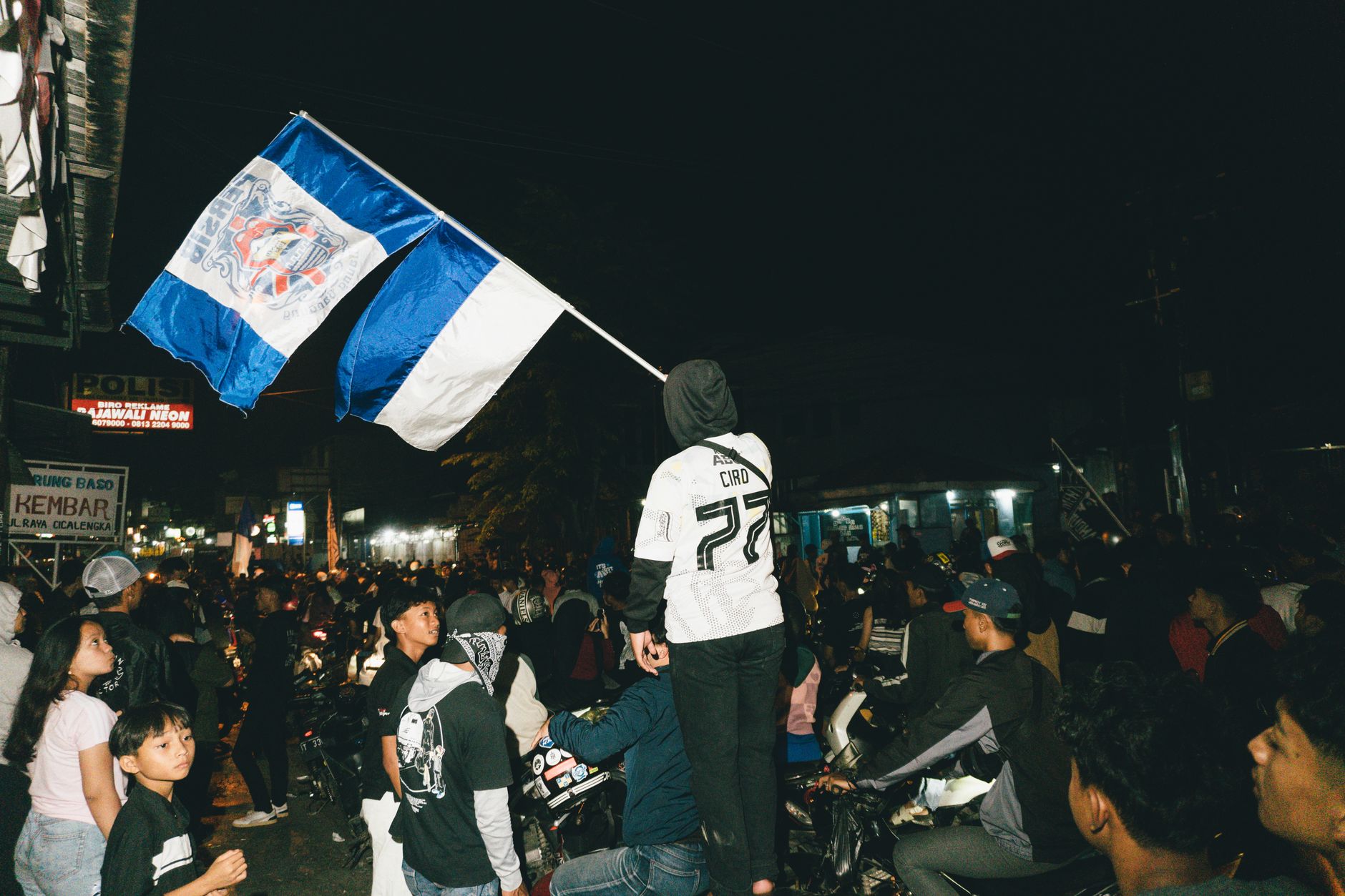 Sebuah kumpulan jalanan yang meriah di malam hari dengan bendera yang ditampilkan secara mencolok di tengah kerumunan besar.