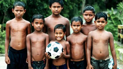 Sekelompok enam anak laki-laki Asia Selatan, berdiri di luar, dengan satu memegang bola sepak.