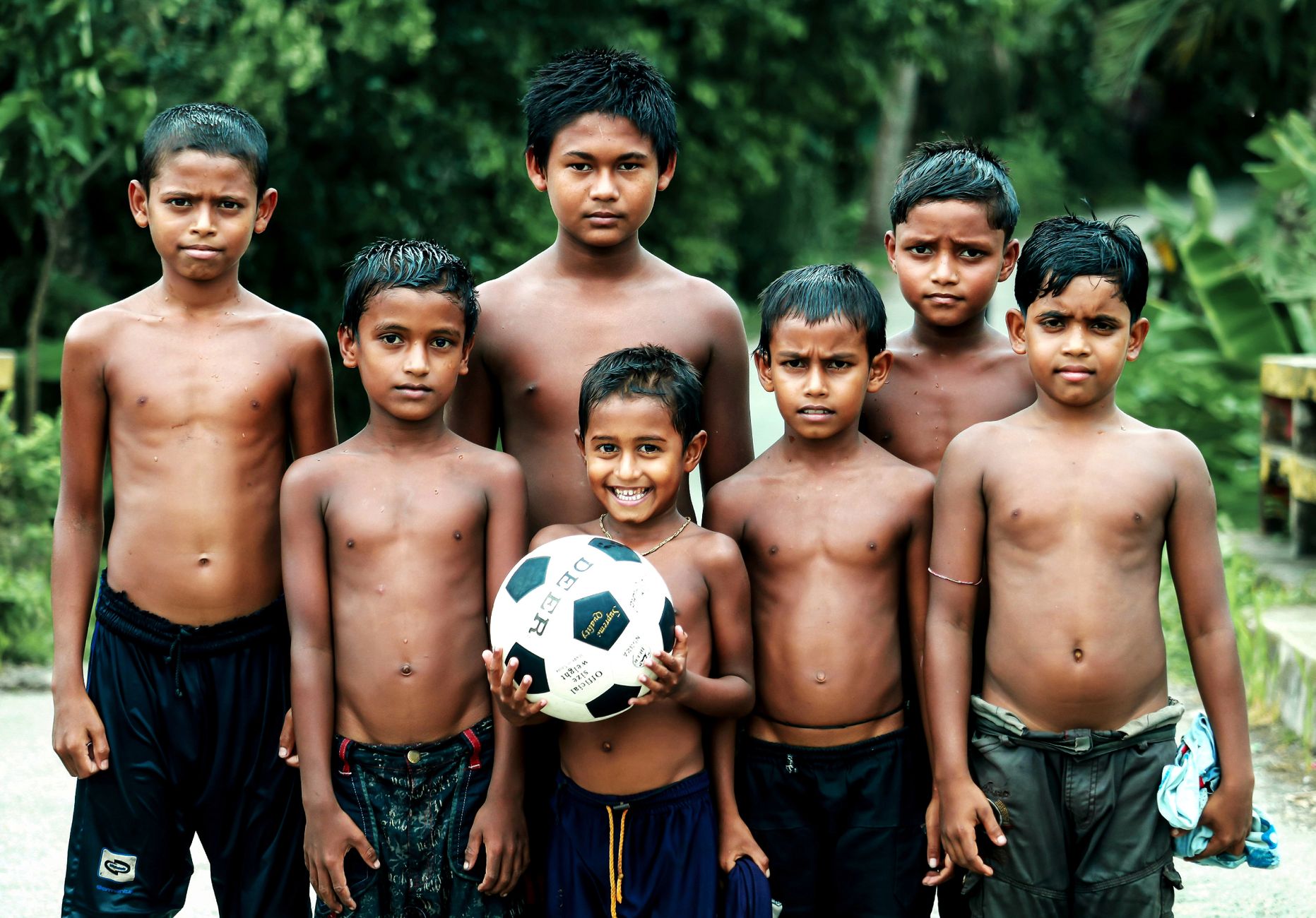Sekelompok enam anak laki-laki Asia Selatan, berdiri di luar, dengan satu memegang bola sepak.