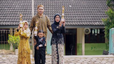 Keluarga yang bangga memegang piala wisuda dengan pakaian tradisional di luar sebuah bangunan.